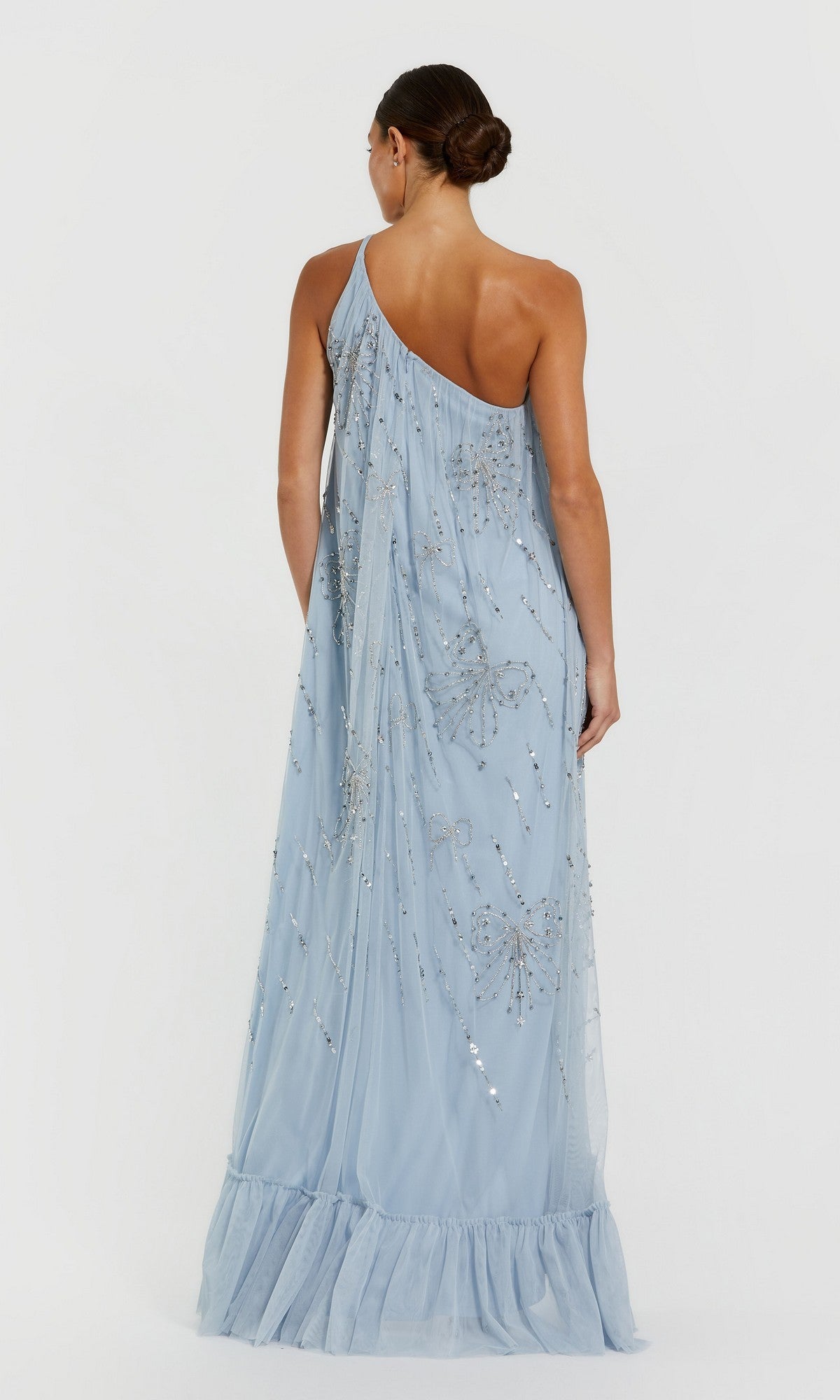 A woman faces away, wearing the Mac Duggal 9344 Long Formal Dress—a light blue, one-shoulder shift with sequin embellishments and a ruffled hem. Her hair is styled in a low bun.